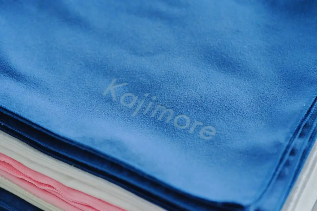 Kajimoreのタオルが3色(ブルー、ピンク、ホワイト)重ねられており、一番上のブルーのタオルにKajimoreのロゴが写っているクローズアップ画像。