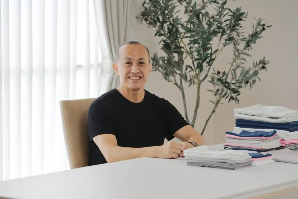 Kajimoreのタオルを前に、優しい笑顔でカメラを見つめている開発者の吉川一平氏の画像。