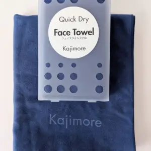 ブルーの速乾フェイスタオルの上に、専用の円形ラベル付きハードケースが置かれた状態。タオル下部には「Kajimore」のロゴが同系色で刻印されている。