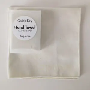 アイボリーの速乾ハンドタオルと、その上に重ねられた専用ハードケース。 タオルの端に「Kajimore」のロゴ刻印が確認できる。